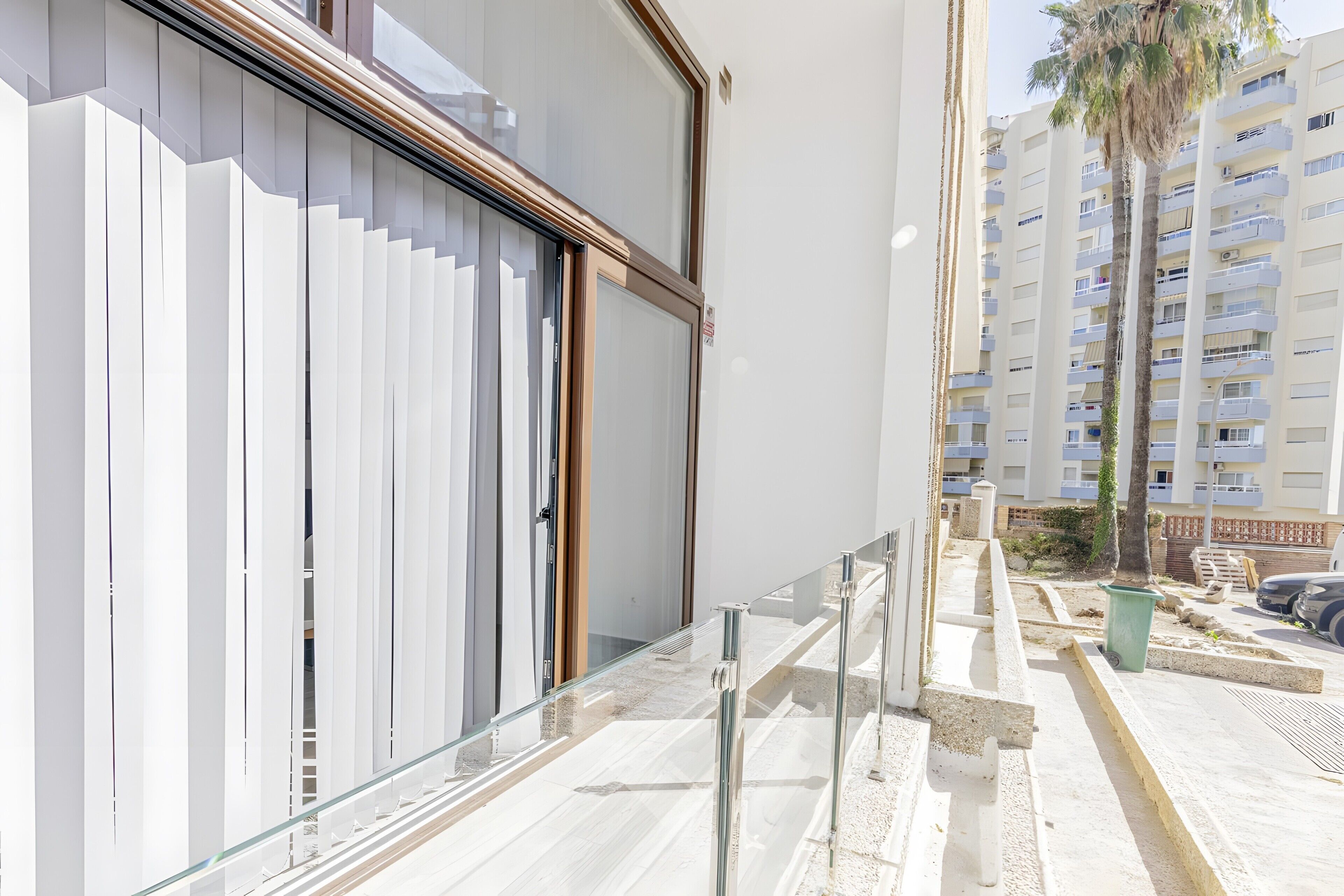 A modern building with a glass facade, a metal railing, and a clear view of a residential area with multi-story buildings.