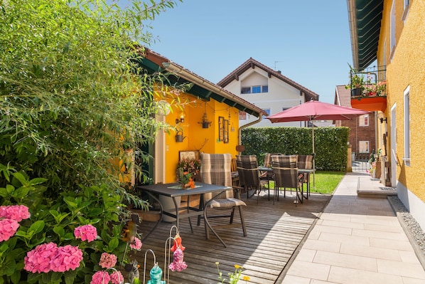 Un patio con mesa y sillas de madera, rodeado de follaje y flores.