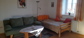 Une petite pièce accueillante, avec un canapé vert, un lit simple agrémenté d’oreillers orange, une petite table sur laquelle se trouve un bocal en verre et une lampe sur pied.