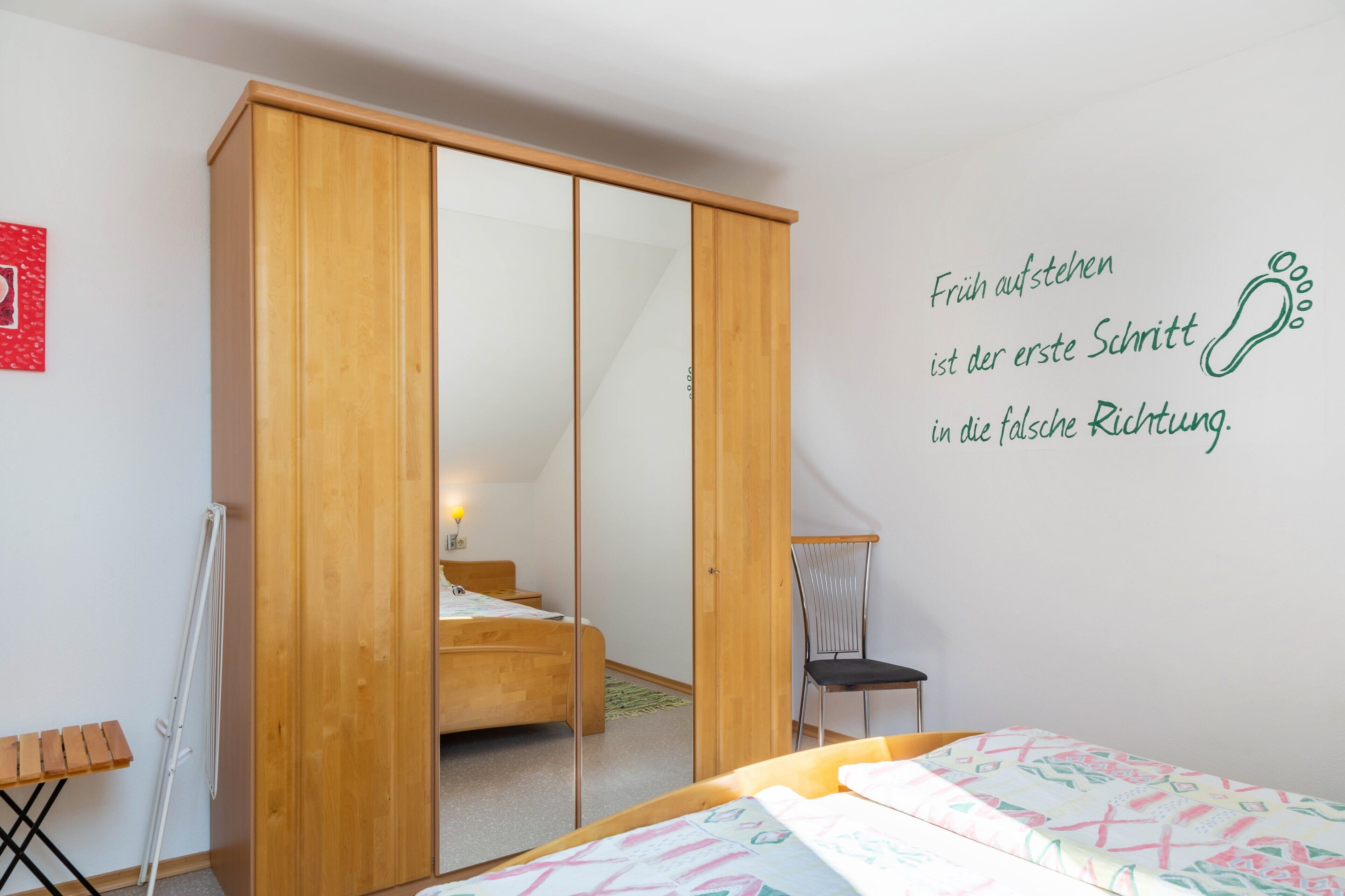 Une chambre avec une armoire en bois, un lit recouvert d’une courtepointe aux couleurs vives, une petite chaise et un mur orné d’une citation inspirante.
