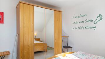 Ein Schlafzimmer mit einem Holzschrank, einem Bett mit farbenfroher Bettdecke, einem kleinen Stuhl und einer Wand mit einem motivierenden Zitat.