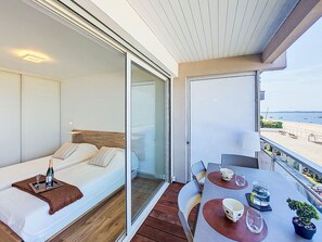 Un balcón con una mesa preparada para dos, una cama con una botella y vasos, y vista a la playa.