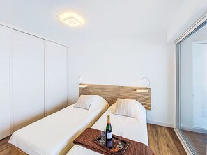 Een moderne hotelkamer met twee bedden, een houten hoofdeinde, een groot raam en een minibar met een fles en glazen.