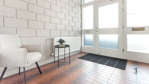 A room with a white brick wall, a white door with frosted glass panels, a white radiator, a black side table with a plant, a white chair, and a black doormat.