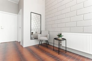 Ein Zimmer mit einer weißen Tür, einem Sessel, einem kleinen Tisch mit Pflanze, einem Spiegel an der Wand und einer weißen Ziegelwand.