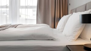 A neatly made bed with white linens, a bedside lamp, and a view of the outdoors through sheer curtains.