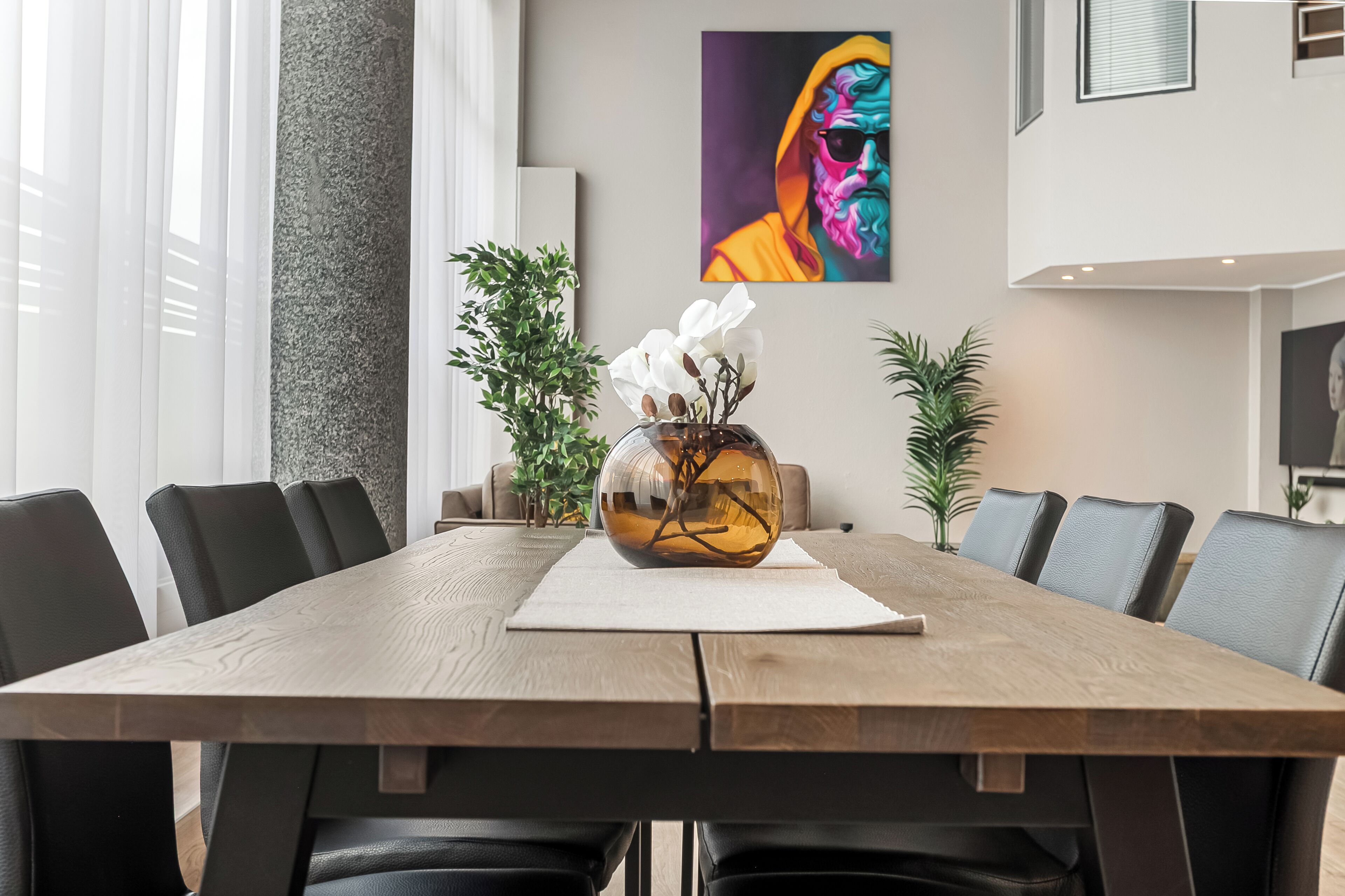 A modern dining area with a wooden table, black chairs, a vase with white flowers, and a colorful painting on the wall.