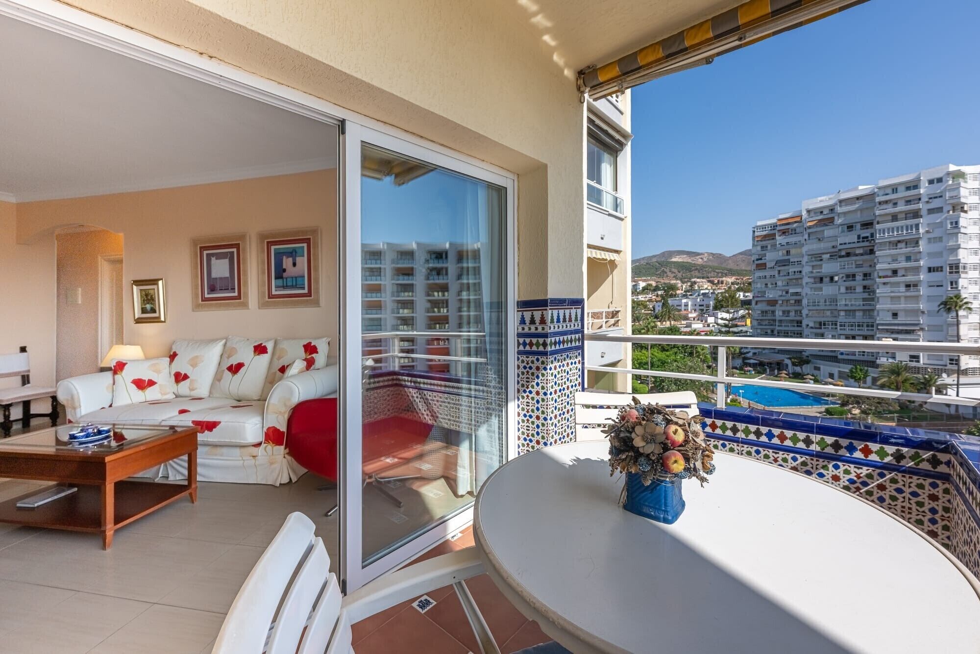 A balcony with a white table, a sofa with red cushions, and a glass door leading to a pool area.