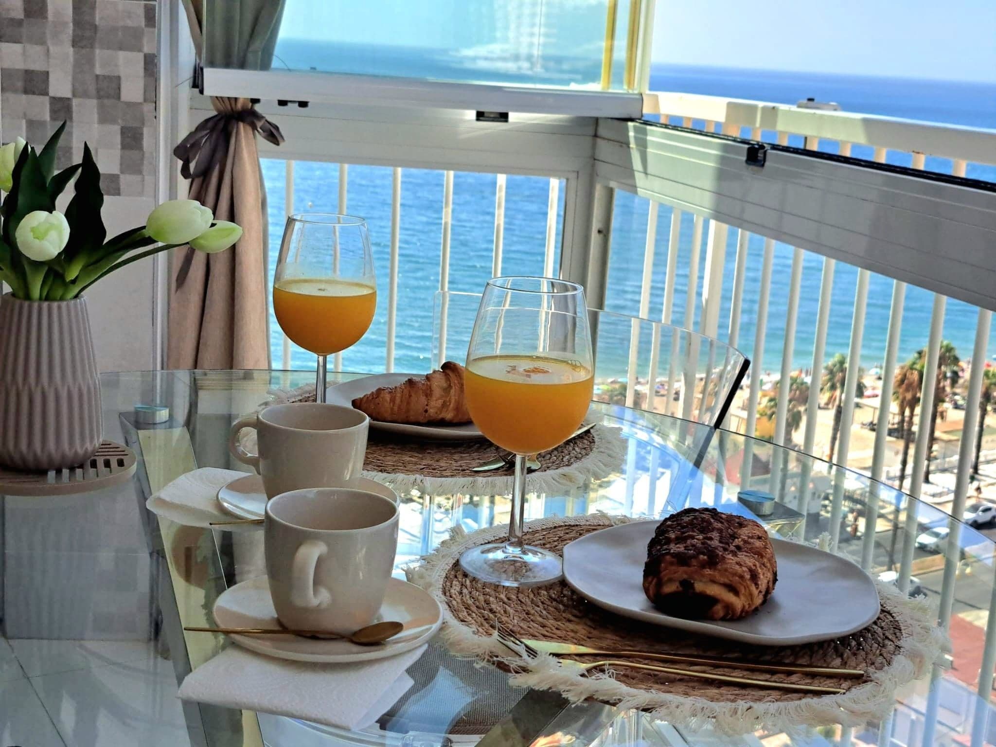 Et bord med appelsinjuice, et croissanter og en kop kaffe, med udsigt over en strand og havet.