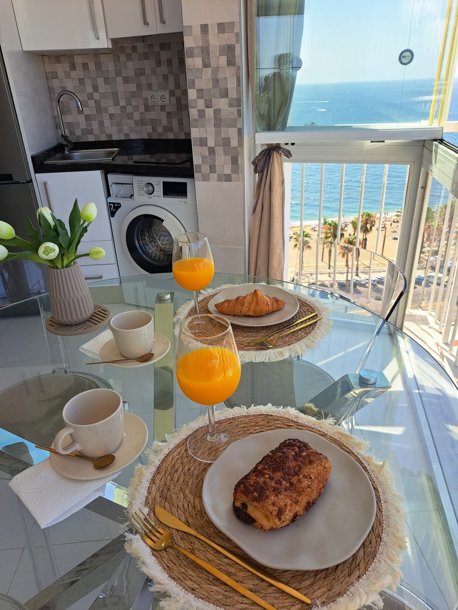 Morgenmad på en balkon med udsigt til havet, med croissanter, appelsinjuice og kaffe.