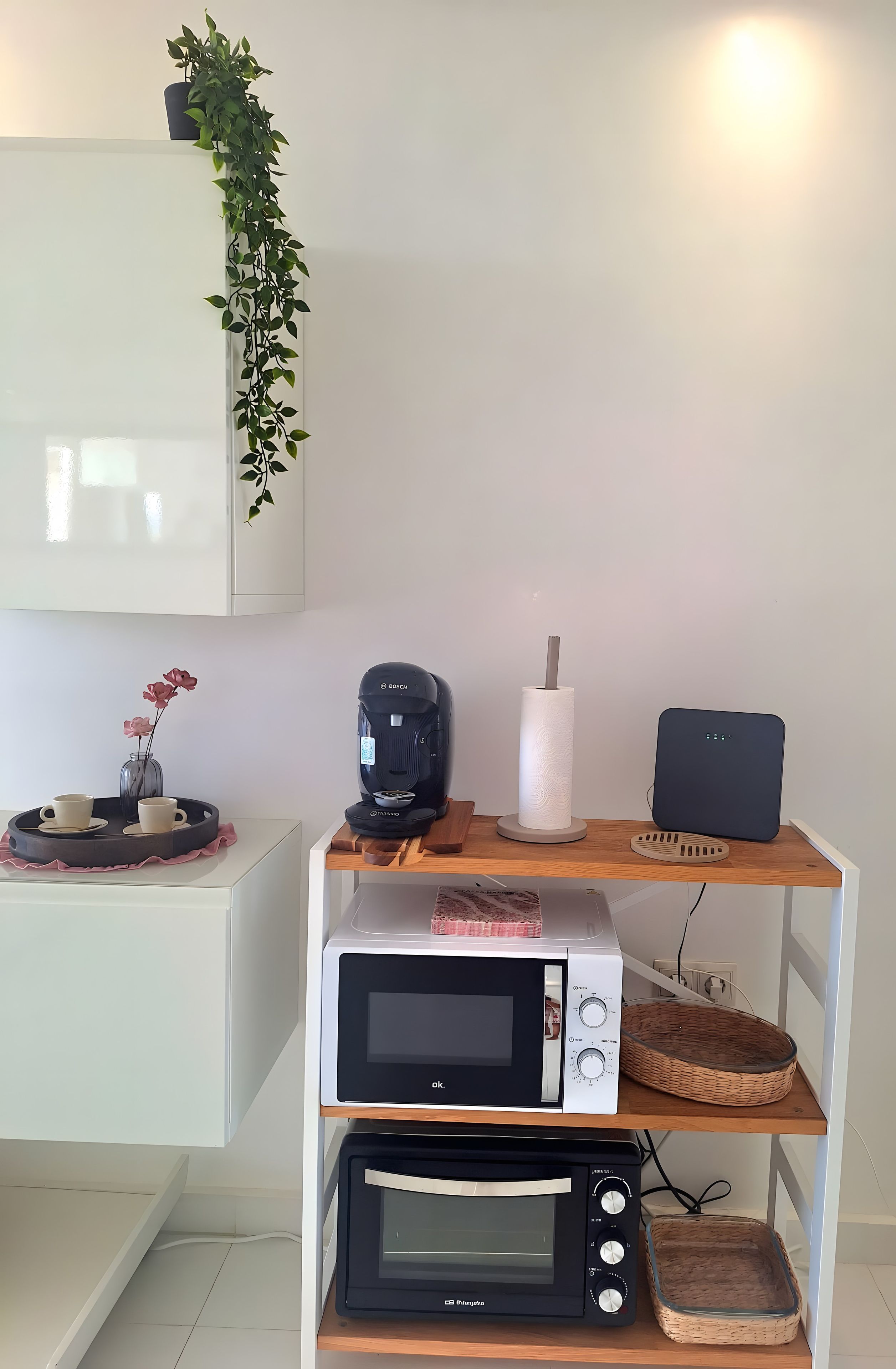 Una cucina con forno a microonde, tostapane e macchina del caffè su un ripiano in legno, una pianta in vaso e un vaso con fiori.