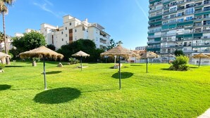 A well-maintained grassy area with thatched umbrellas, a multi-story building in the background, and a clear sky. - Beachfront, 12th Floor with Stunning Views, Maite 2, Benalmádena (Benalmádena)