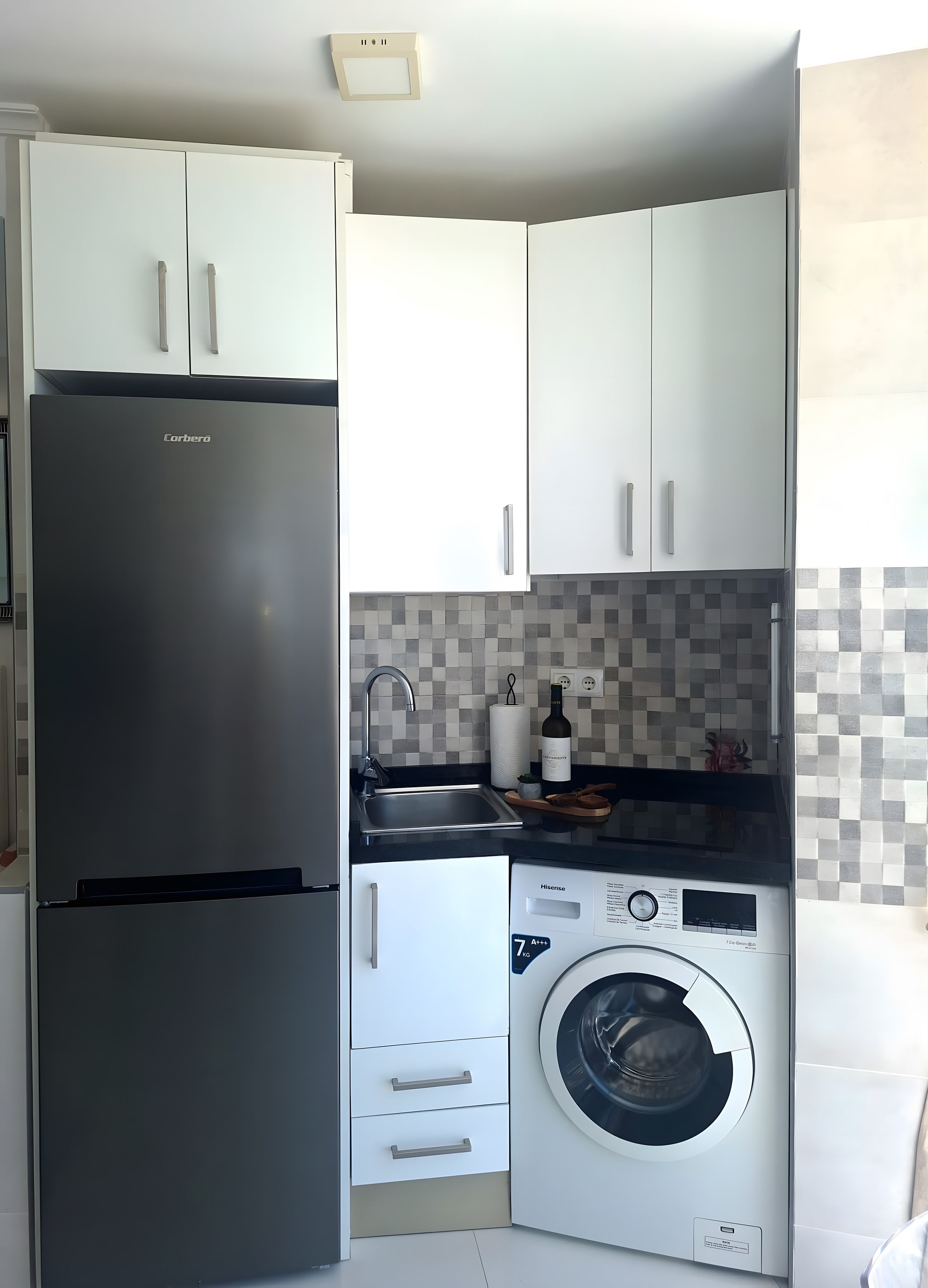 Une cuisine compacte dotée d’un réfrigérateur en acier inoxydable, d’un lave-linge et d’un dosseret carrelé.
