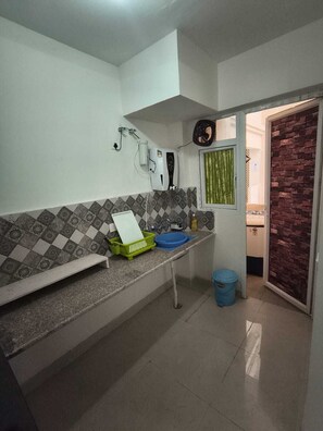 A kitchen with a countertop, a window with blinds, a brick wall, and a door leading to another room. - Stylish 2 Bedroom Apartment in Gurugram (Gurugram)