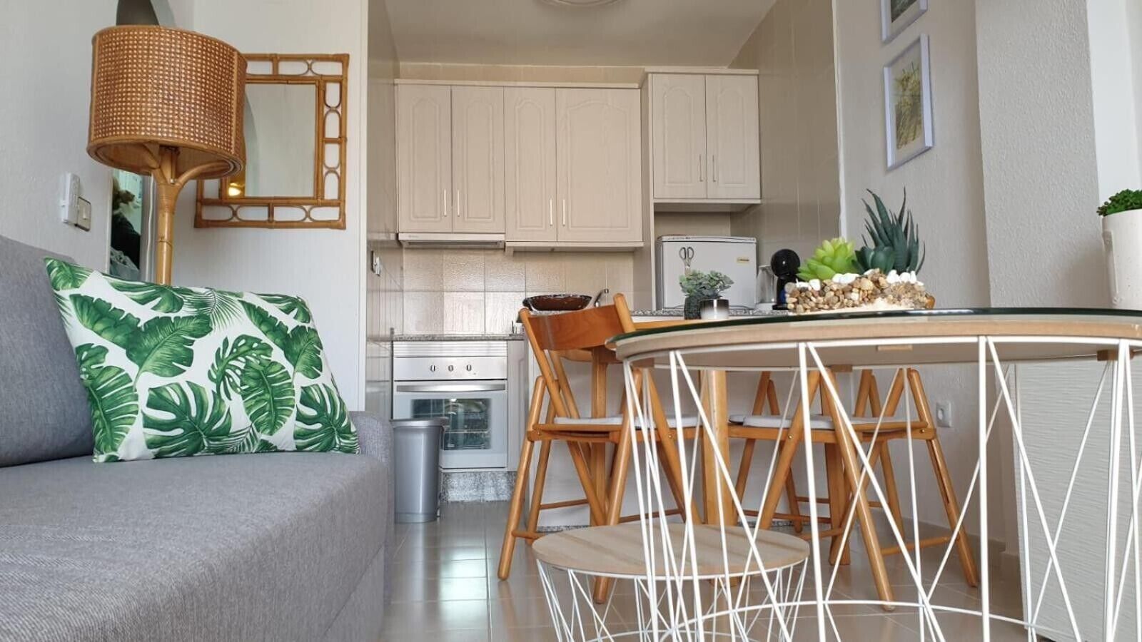 Una cocina moderna con armarios blancos, una mesa de comedor con sillas, un sofá con un cojín estampado tropical y una lámpara de mimbre.