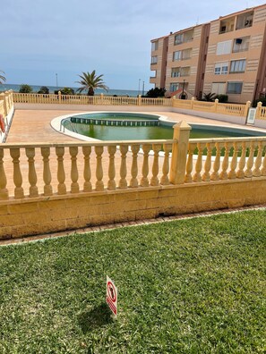 A swimming pool surrounded by a fence, a grassy area, and a building with balconies.