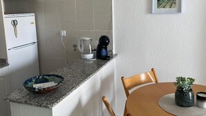 A compact kitchen with a refrigerator, coffee maker, and a small island with a bowl. There is a dining table with chairs and a potted plant.