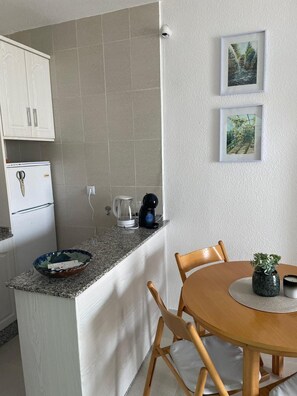 Una cocina compacta con refrigerador, cafetera y una pequeña isla con un tazón. Hay una mesa de comedor con sillas y una planta en maceta.