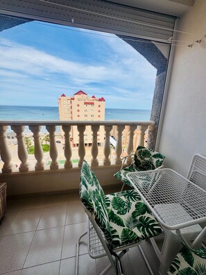 A balcony with a table and chairs, a view of the sea, and a building in the background.