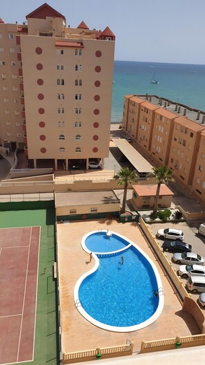 A multi-story hotel with a swimming pool, tennis court, and a view of the sea.
