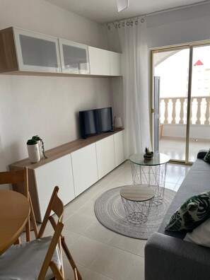 Un espacio habitable moderno con sofá, mesa de comedor, televisor en un estante y balcón con barandilla.