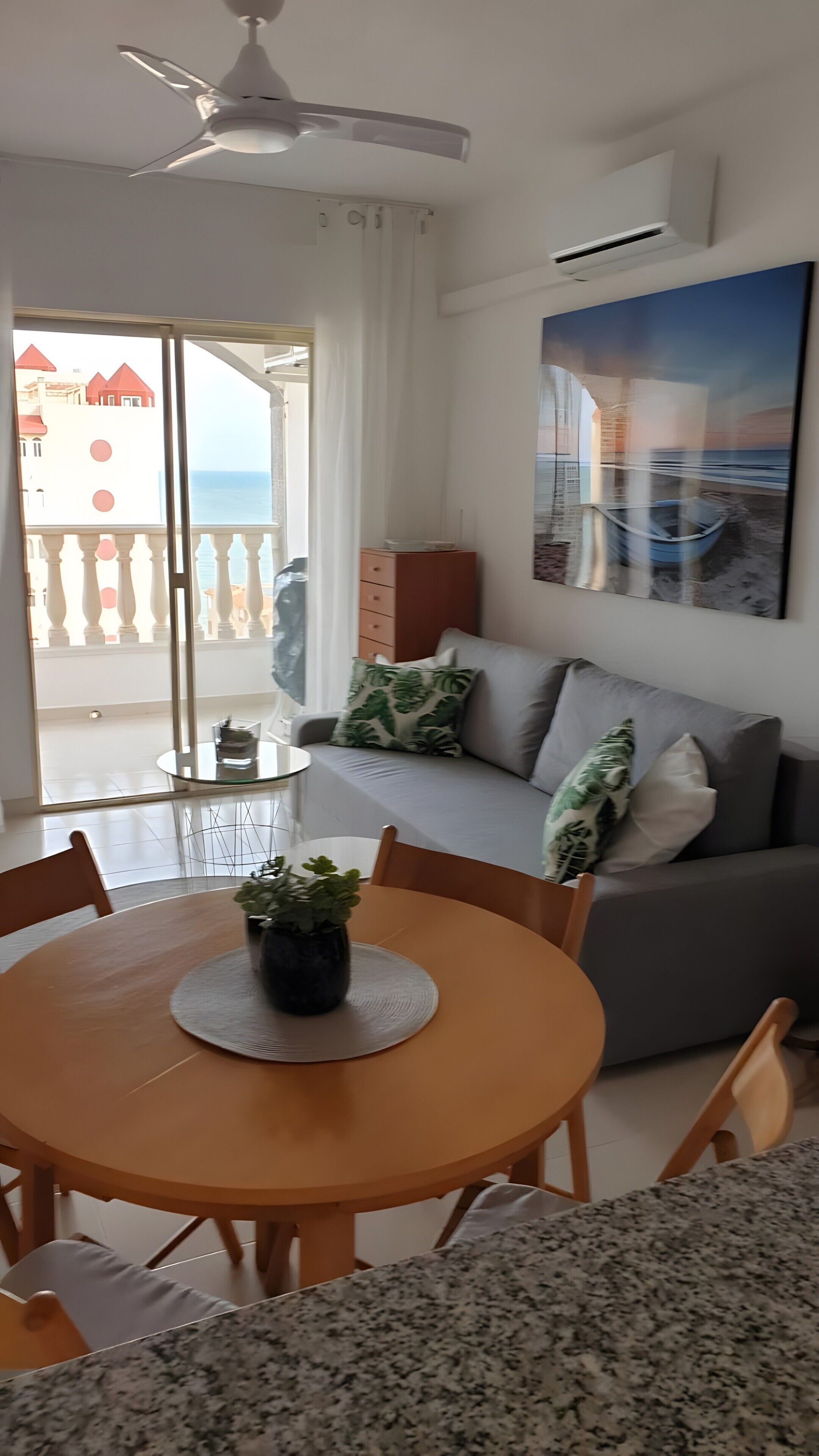 Ein Essbereich mit rundem Tisch, Stühlen, einer Zimmerpflanze, einer Couch mit Kissen, einem Deckenventilator und einem großen Fenster mit Blick auf Gebäude.