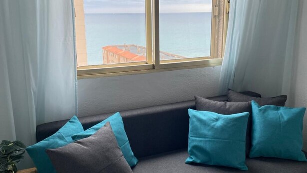 Ein graues Sofa mit türkisfarbenen Kissen, ein kleiner Tisch mit einer Pflanze und ein Fenster mit Blick auf das Meer und Gebäude.