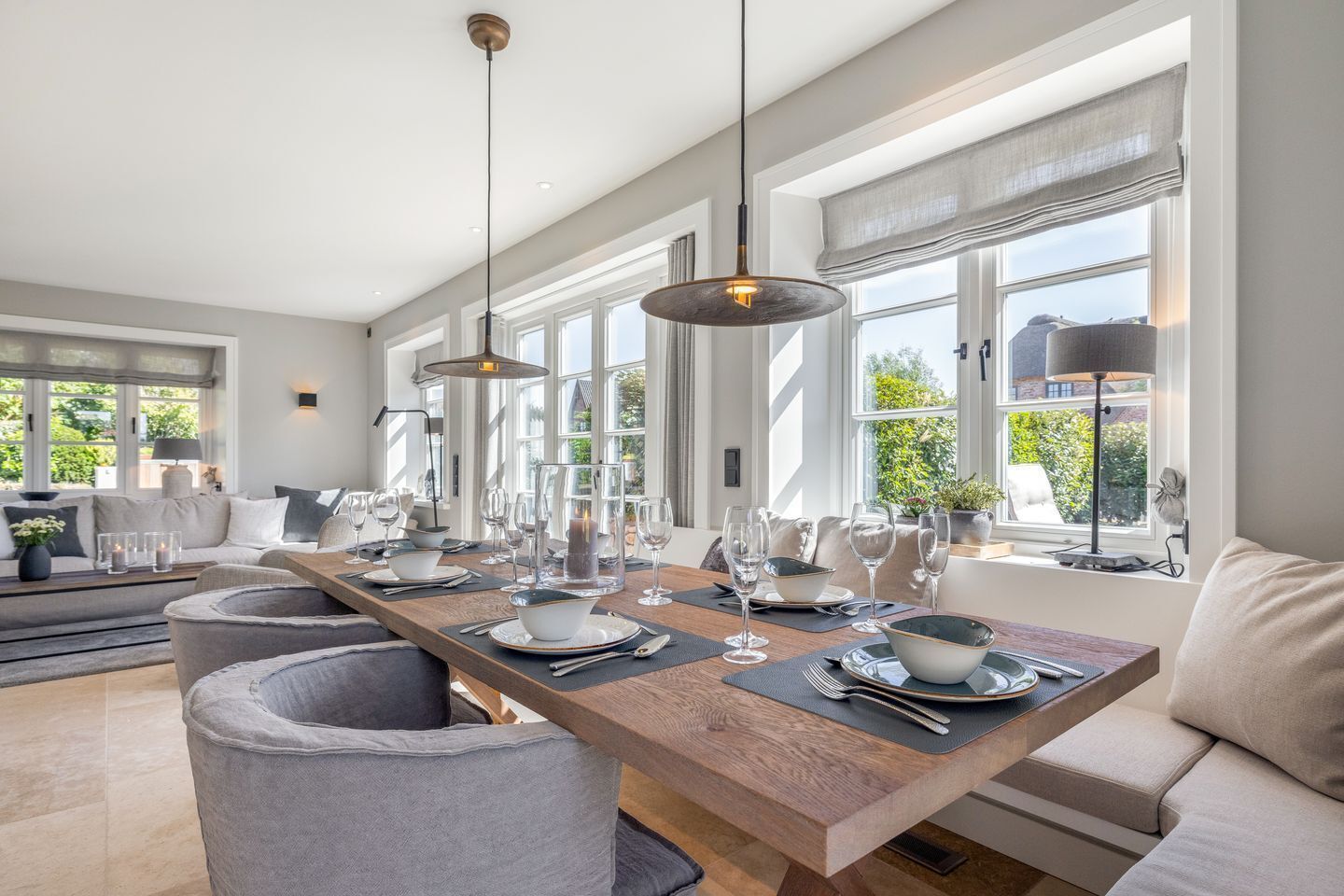A modern dining area with a long wooden table set for six, large windows, and a cozy seating area with a sofa and armchairs.