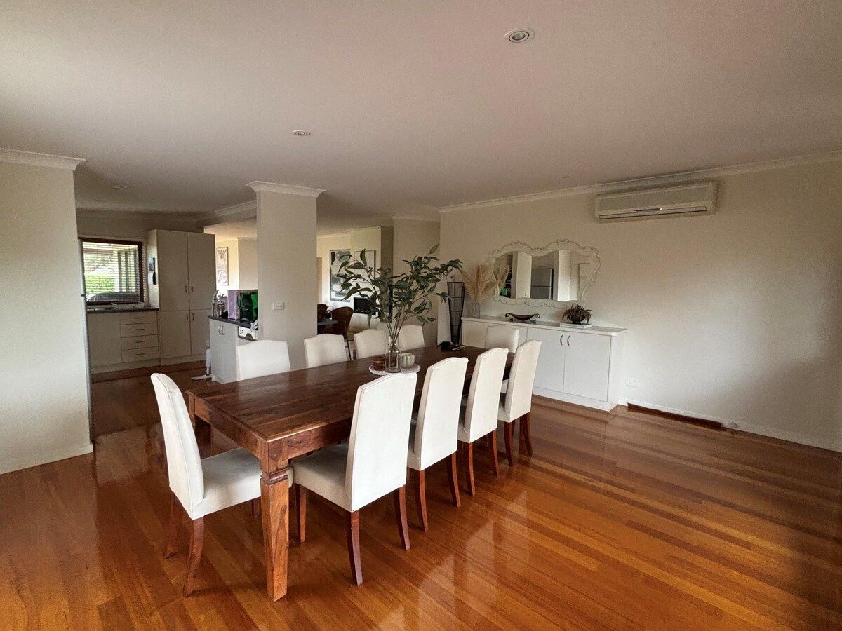 Uma área de refeições com mesa de madeira e cadeiras brancas, uma cozinha ao fundo e uma parede branca com um aparelho de ar condicionado.