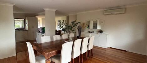 Un coin repas avec une table en bois et des chaises blanches, une cuisine en arrière-plan et un mur blanc avec un appareil de climatisation.