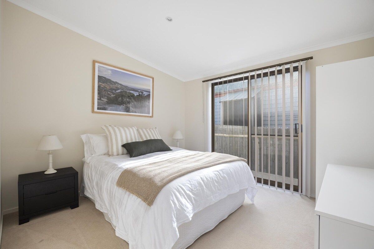 A bedroom with a bed, a nightstand, a window with blinds, and a framed picture on the wall.