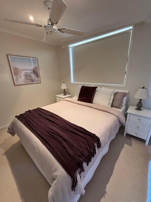 Een slaapkamer met een bed, nachtkastjes, een plafondventilator, een raam met jaloezieën en een lijst met een afbeelding aan de muur.