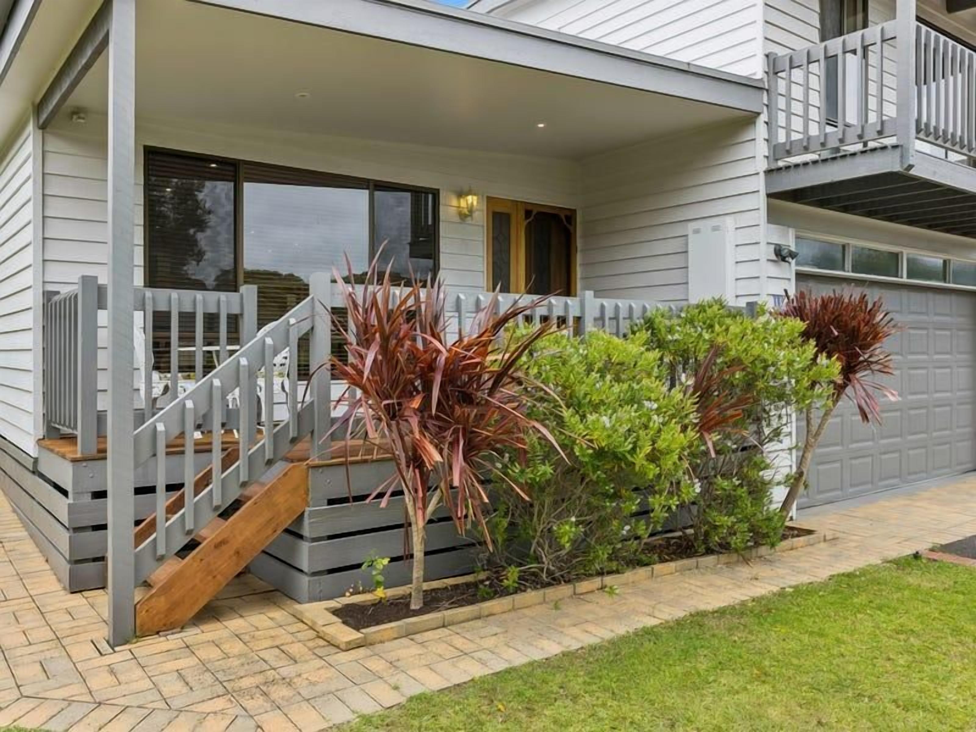 A modern two-story house with a covered porch, a wooden staircase, and a small garden with red-leaved plants.