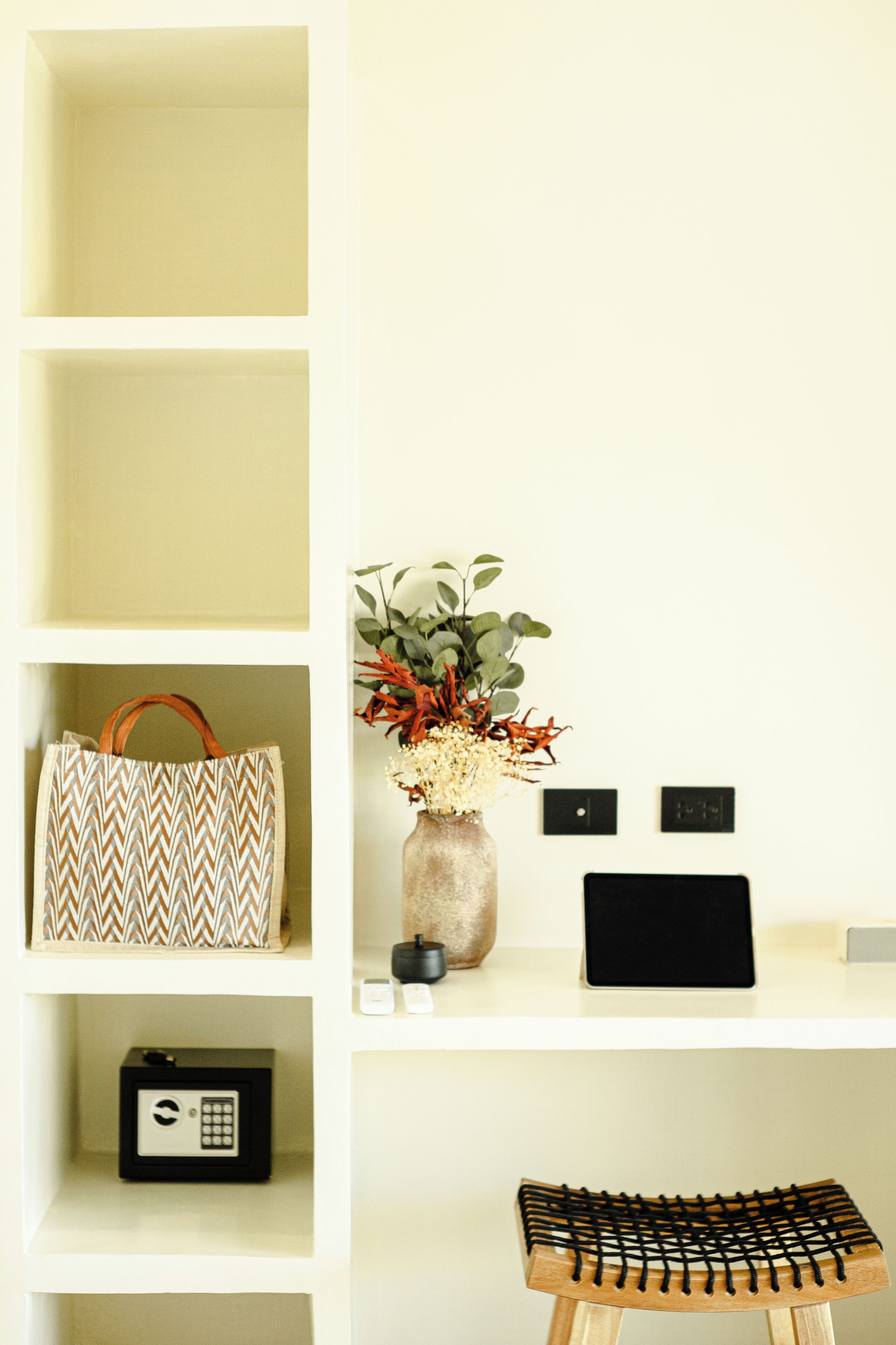 A shelf with a patterned bag, a vase with flowers, a small black box, and a wooden stool.