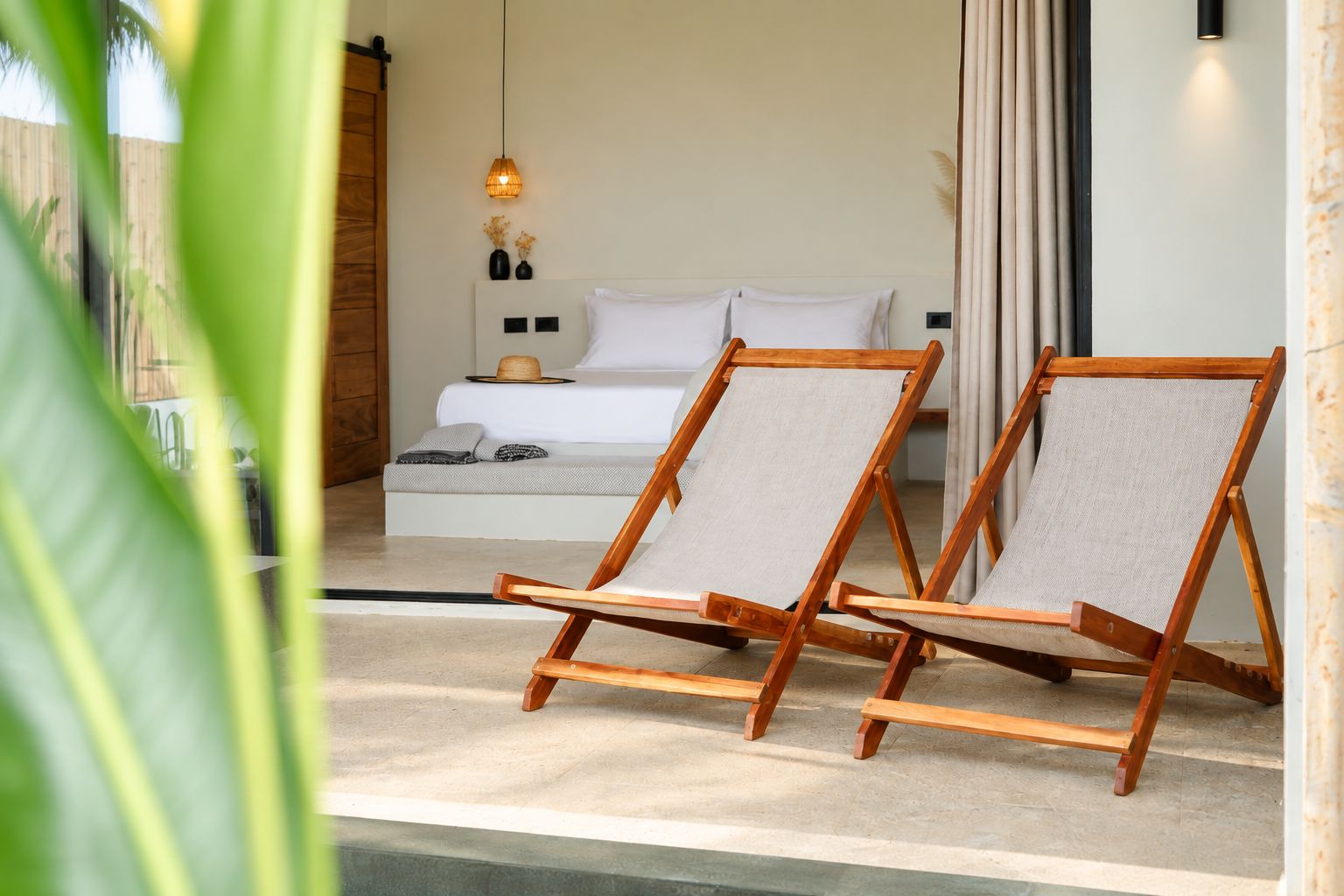 A bedroom with a bed, two wooden chairs, a bedside table, a wall-mounted lamp, and a view of the outdoors.