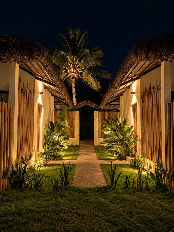 A pathway leading to a building with a thatched roof and bamboo walls, surrounded by tropical plants.