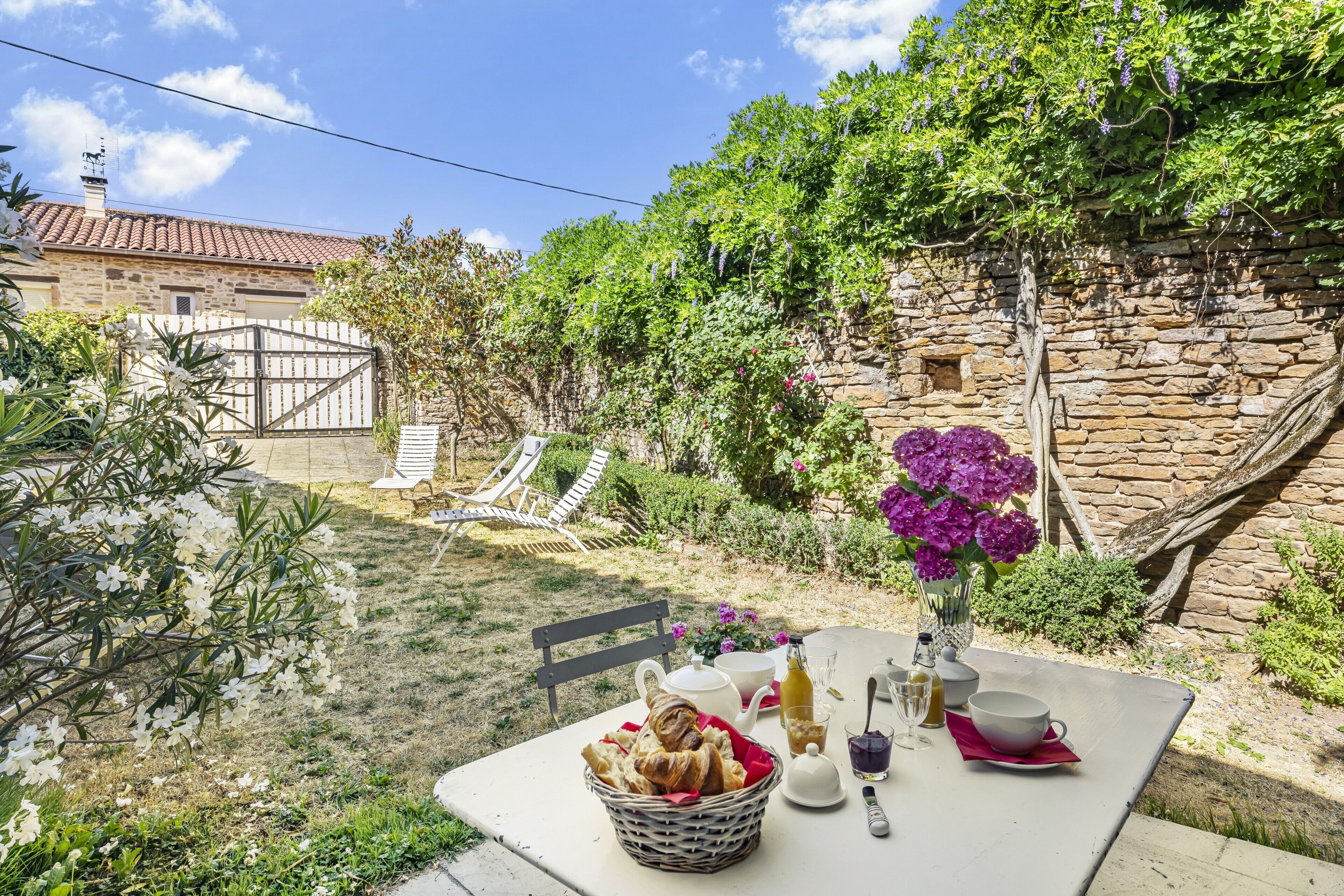 An outdoor dining area with a table set for breakfast, including bread, a teapot, cups, and a vase with flowers.