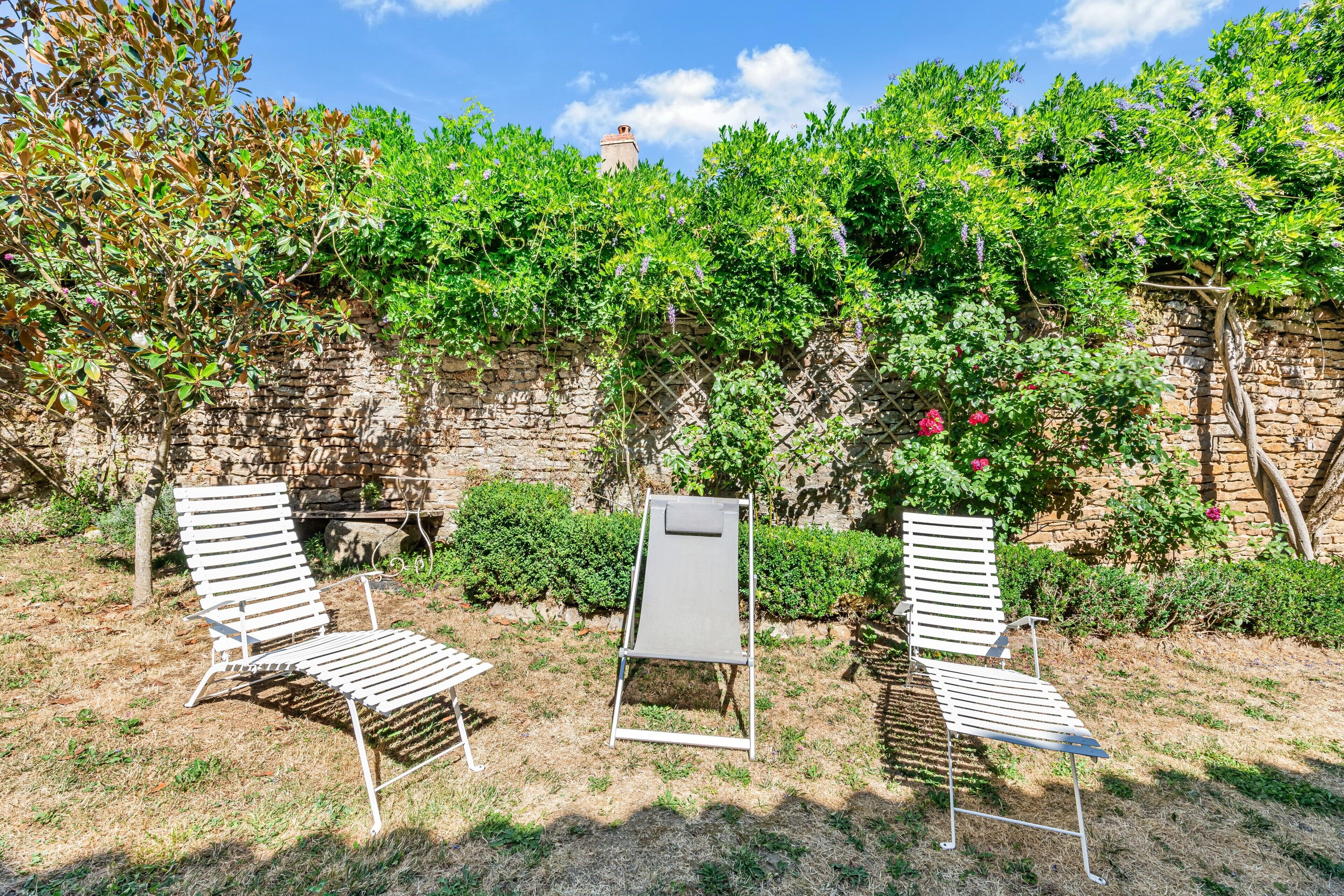 Zwei weiße Metall-Loungesessel in einem Garten mit Steinmauer und Grünpflanzen im Hintergrund.