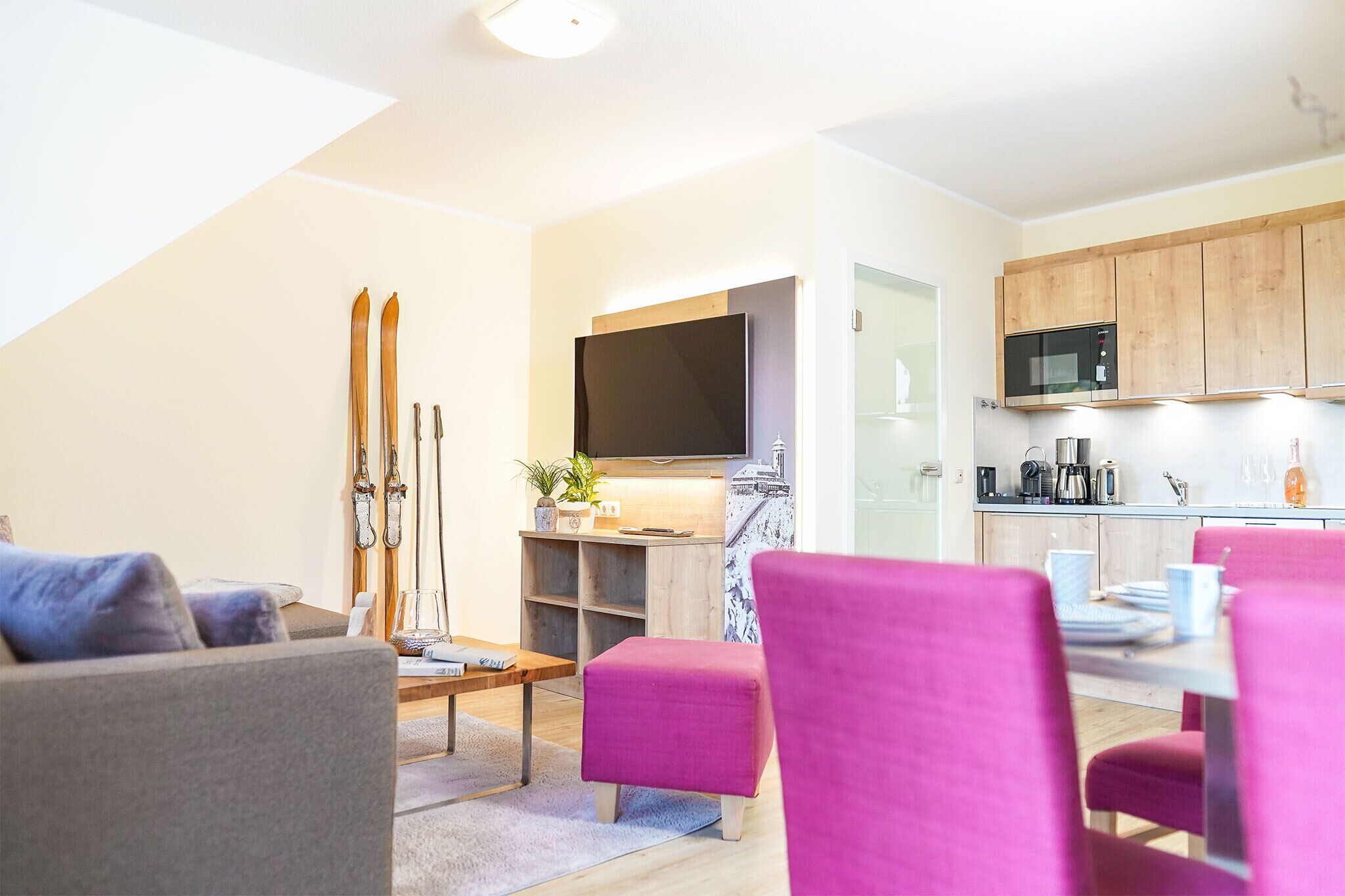 A modern living room with a sofa, a coffee table, and a dining area with chairs. There is a kitchen with wooden cabinets and a microwave.