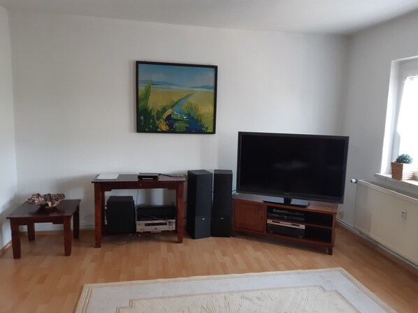 A living room with a flat-screen TV, a wooden entertainment unit, a painting on the wall, and a window with a plant.