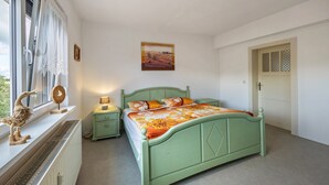 Un dormitorio con cama verde, una mesita de noche, lámpara, radiador, ventana con cortinas y puerta con panel decorado.