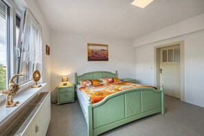 A bedroom with a green bed, a nightstand, a lamp, a radiator, a window with curtains, and a door with a patterned panel.