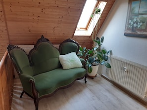 A green upholstered sofa with a white cushion, a potted plant, and a radiator in a room with wooden walls and a sloped ceiling.