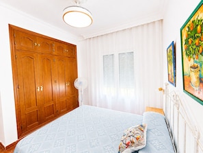 Ein Schlafzimmer mit großem Holzschrank, einem Bett mit gemusterten Bettwaren, einem Standventilator und einem Fenster mit Jalousien.
