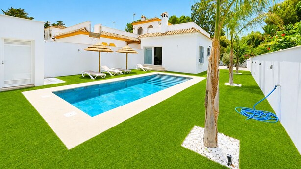 Una piscina con sillas reclinables blancas, una palmera y un edificio blanco con techo de tejas.