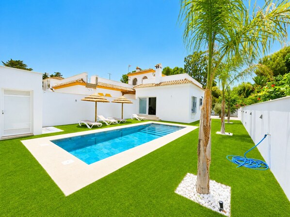 A swimming pool with white lounge chairs, a palm tree, and a white building with a tiled roof.