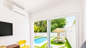 Un salón con televisor, una mesita, un sofá y un ventanal corredizo que da a una zona de piscina con césped y una pérgola.