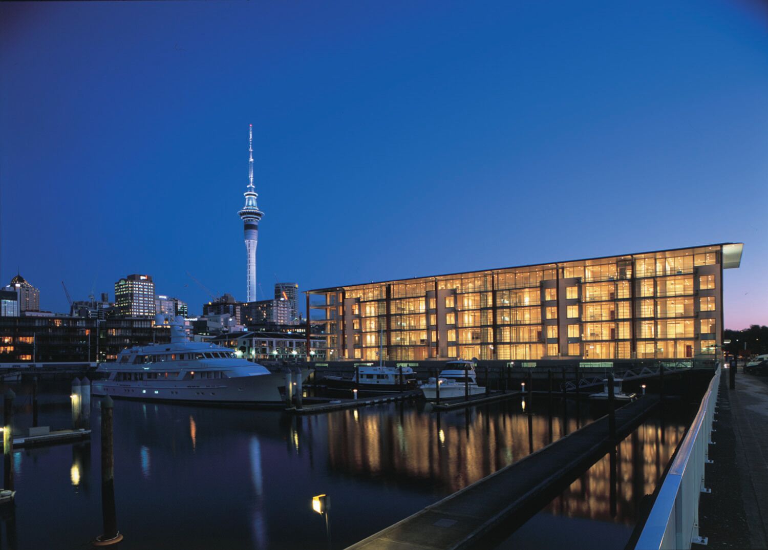 A modern building with a glass facade, a waterfront, and a prominent tower in the background.