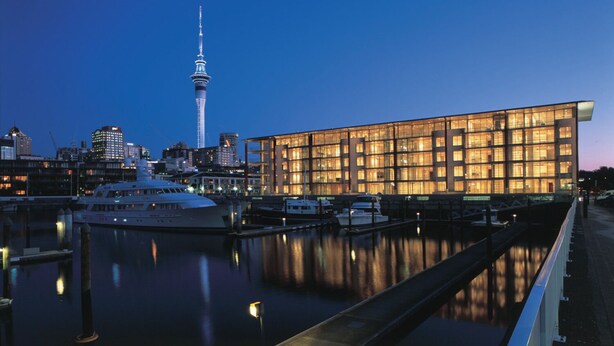 Een modern gebouw met een glazen gevel, een ligging aan het water en een opvallende toren op de achtergrond.