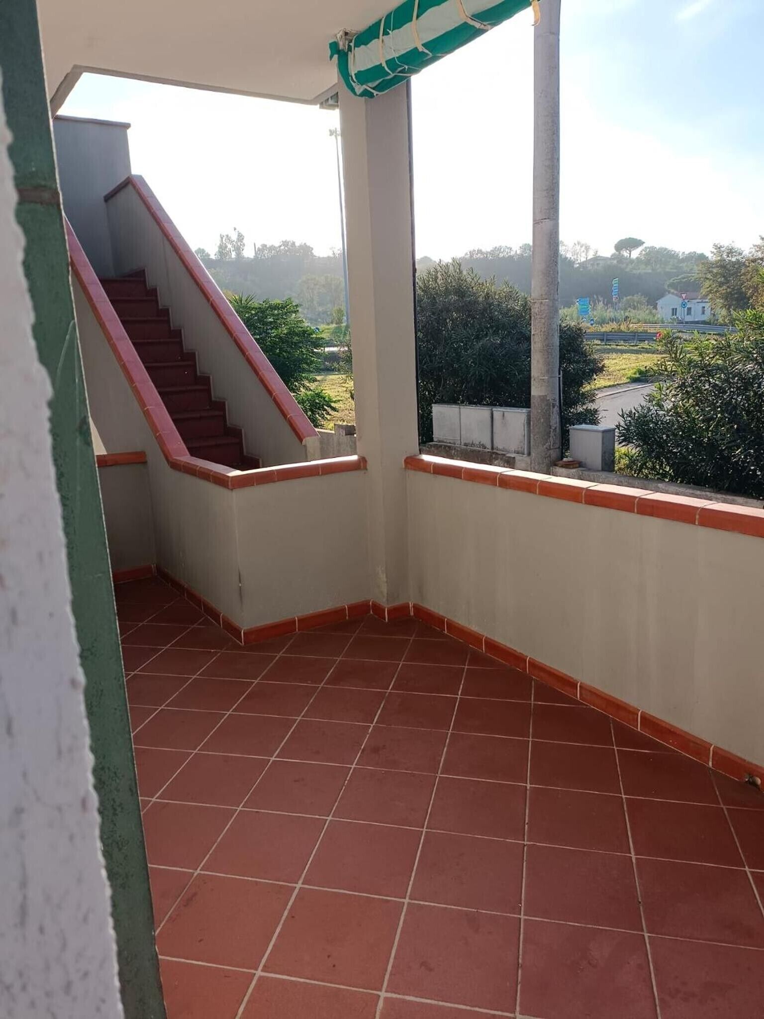 A covered balcony with red-tiled flooring, a staircase, and a view of greenery and buildings in the distance.